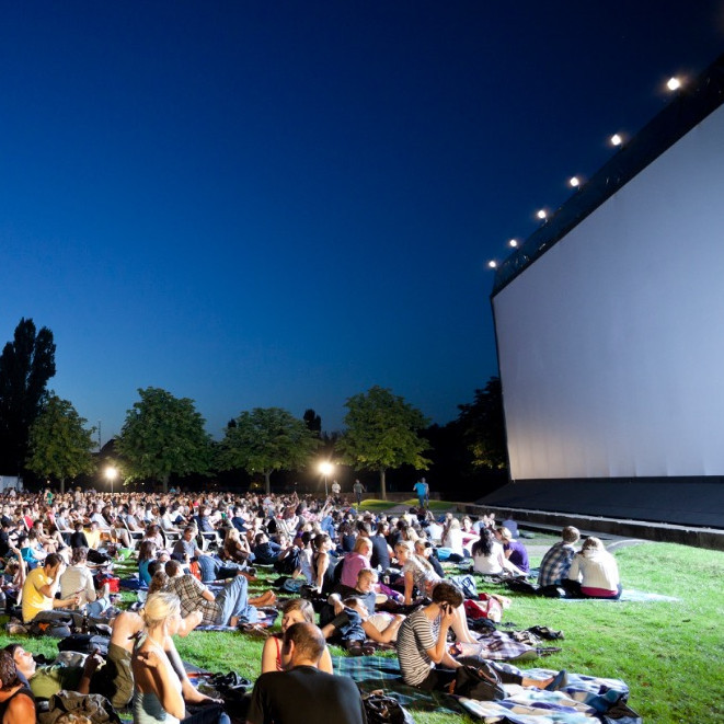 Menschen sitzen auf Wiese vor großer Leinwand.